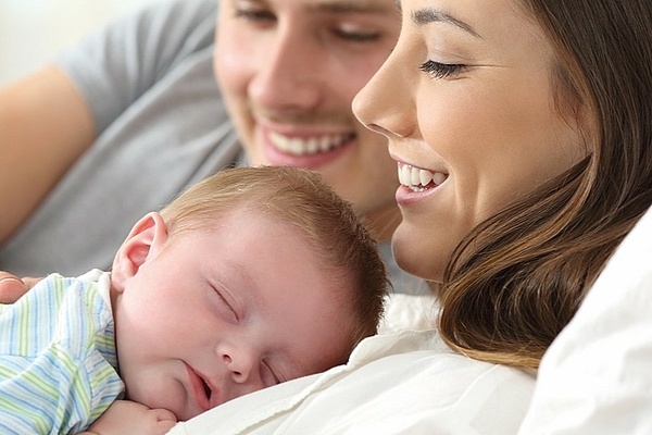 Mutter liegt mit einem Neugeborenen auf dem Bauch im Bett während der Vater danebenliegt, sich an die Frau schmiegt und das Baby am Rücken streichelt