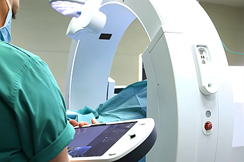 Ein Mediziner in grüner OP-Kleidung und mit blauem Mund-Nase-Schutz bedient über eine digitale Konsole ein medizinisches Gerät für eine hochmoderne Bildgebung. Im Hintergrund ist ein OP-Tisch auf dem ein blaues Tuch ausgebreitet ist.