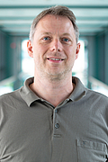 Ein Mann in einem grauen Poloshirt steht in einem lichtdurchfluteten Korridor und lächelt in die Kamera.