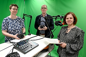 Zwei Frauen und ein Mann stehen in einem Tonstudio an einem Tisch, auf dem ein Mischpult zu sehen ist. Der Hintergrund ist grün. Zwei Personen halten Kopfhörer in der Hand und stehen vor Mikrofonen an Mikrofonarmen. Eine Person hält ein Blatt Papier. 